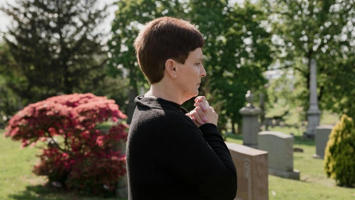 A woman dressed in black stands solemnly in a peaceful cemetery, reflecting amidst gravestones.
