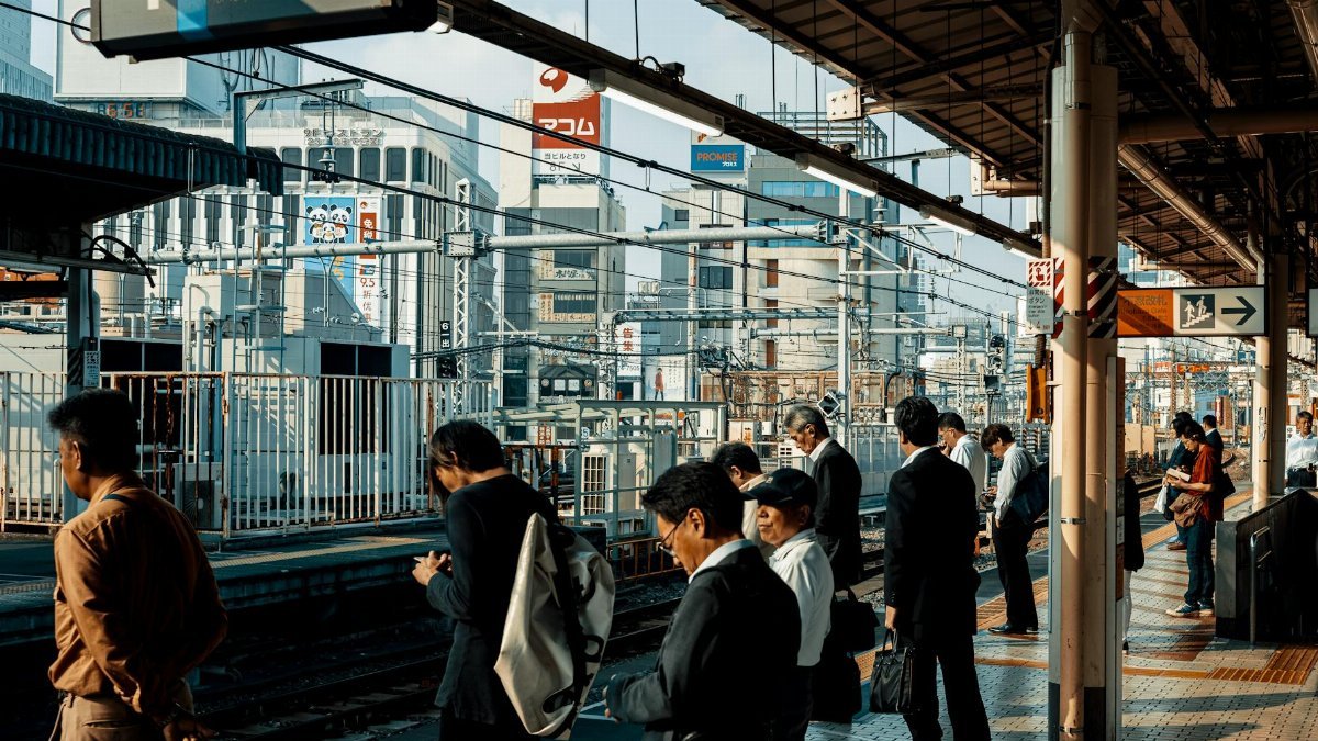 Commuters waiting at a bustling urban train station platform under a modern cityscape.