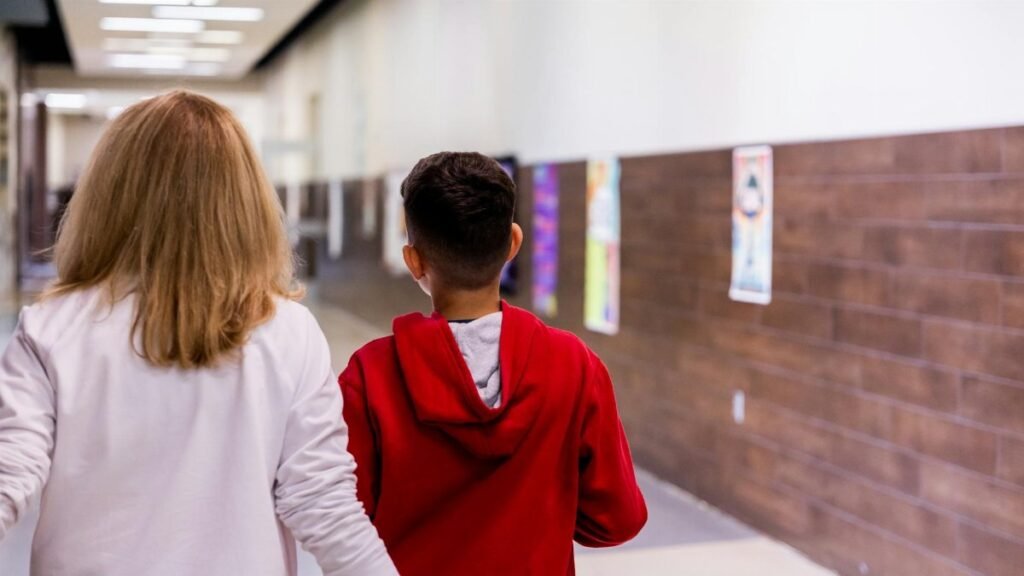 A teacher and student walking together down a brightly lit school hallway.