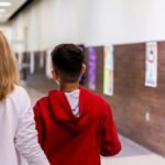 A teacher and student walking together down a brightly lit school hallway.