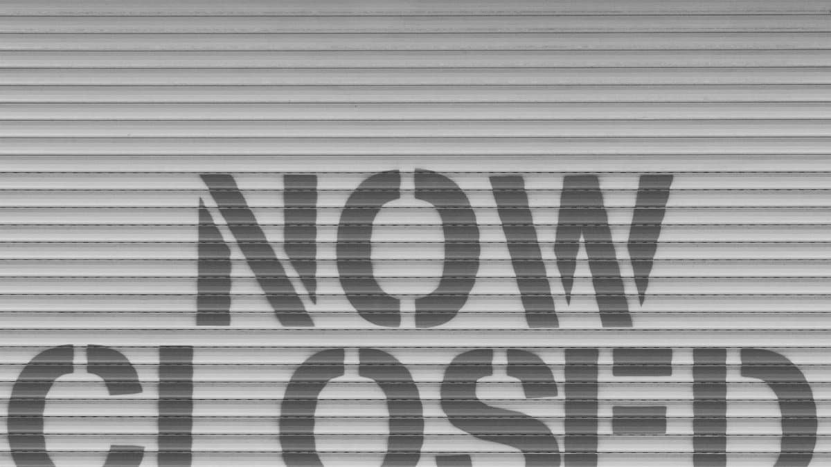 Black and white image of a 'Now Closed' sign on a steel shutter, symbolizing closure.