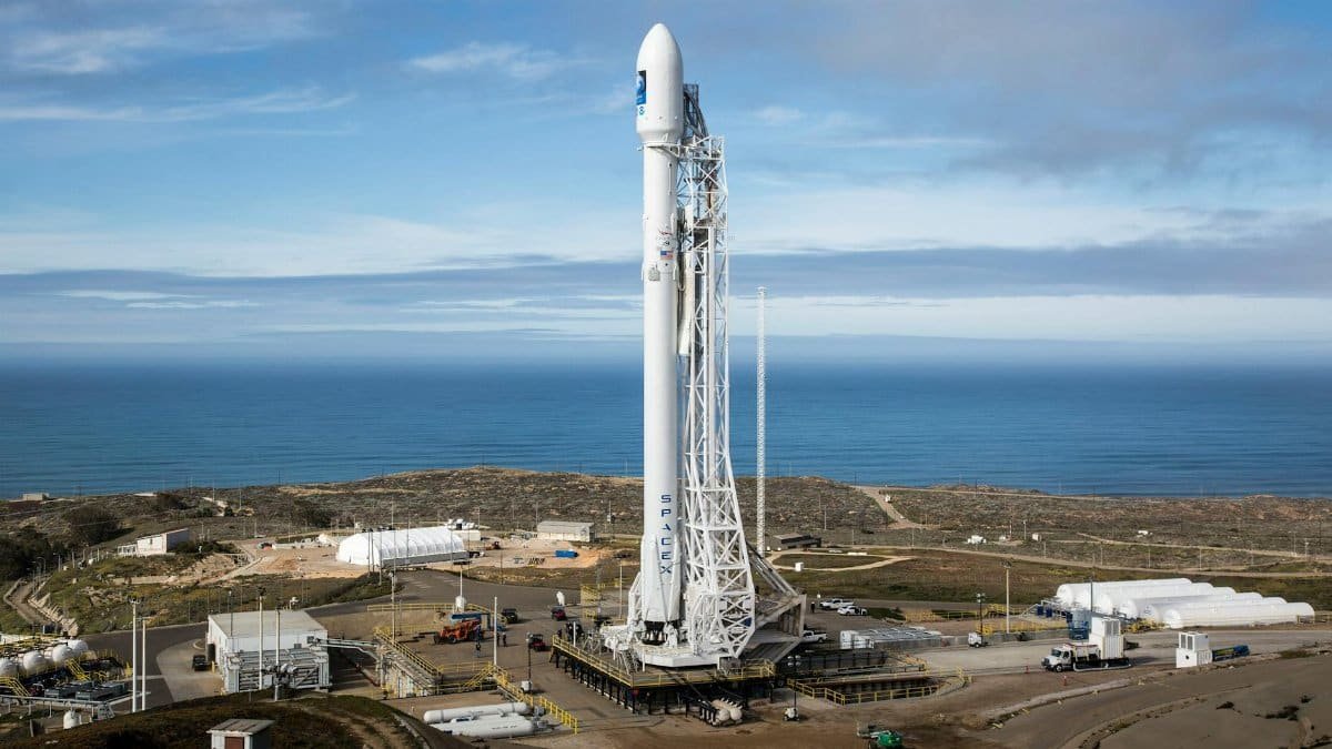 A SpaceX rocket stands on a launch pad overlooking the ocean under a clear sky.