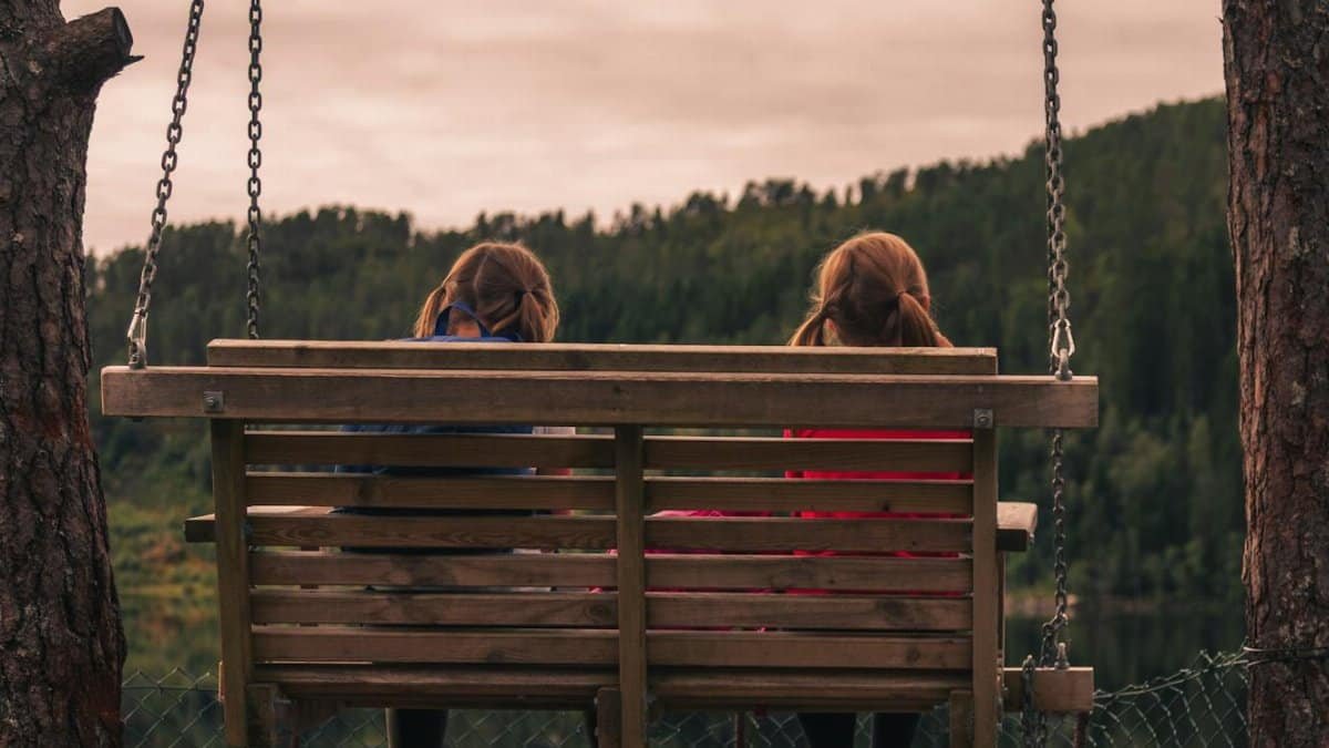 Two children swing on a lakeside bench swing surrounded by trees in a serene Norwegian landscape.