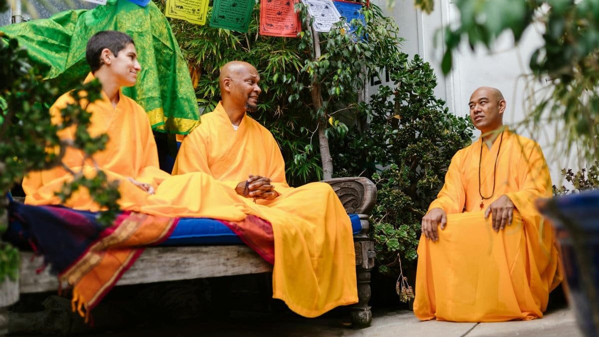 Three monks in bright robes sit in a garden, engaging in a peaceful conversation, surrounded by greenery and prayer flags.