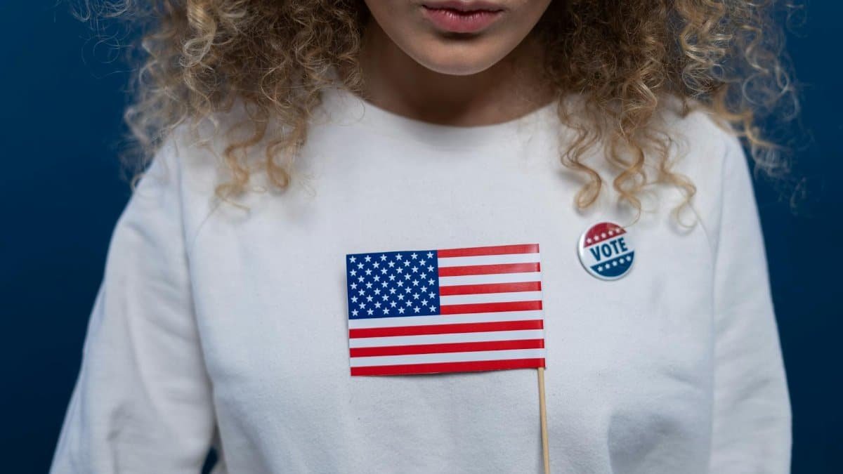 A woman with curly hair holding an American flag and wearing a vote pin against a blue background.
