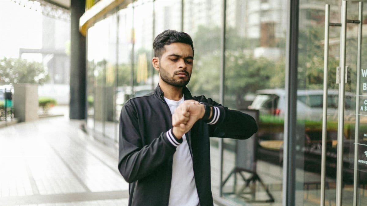 A man in a city looks at his wristwatch while standing near a modern glass building.