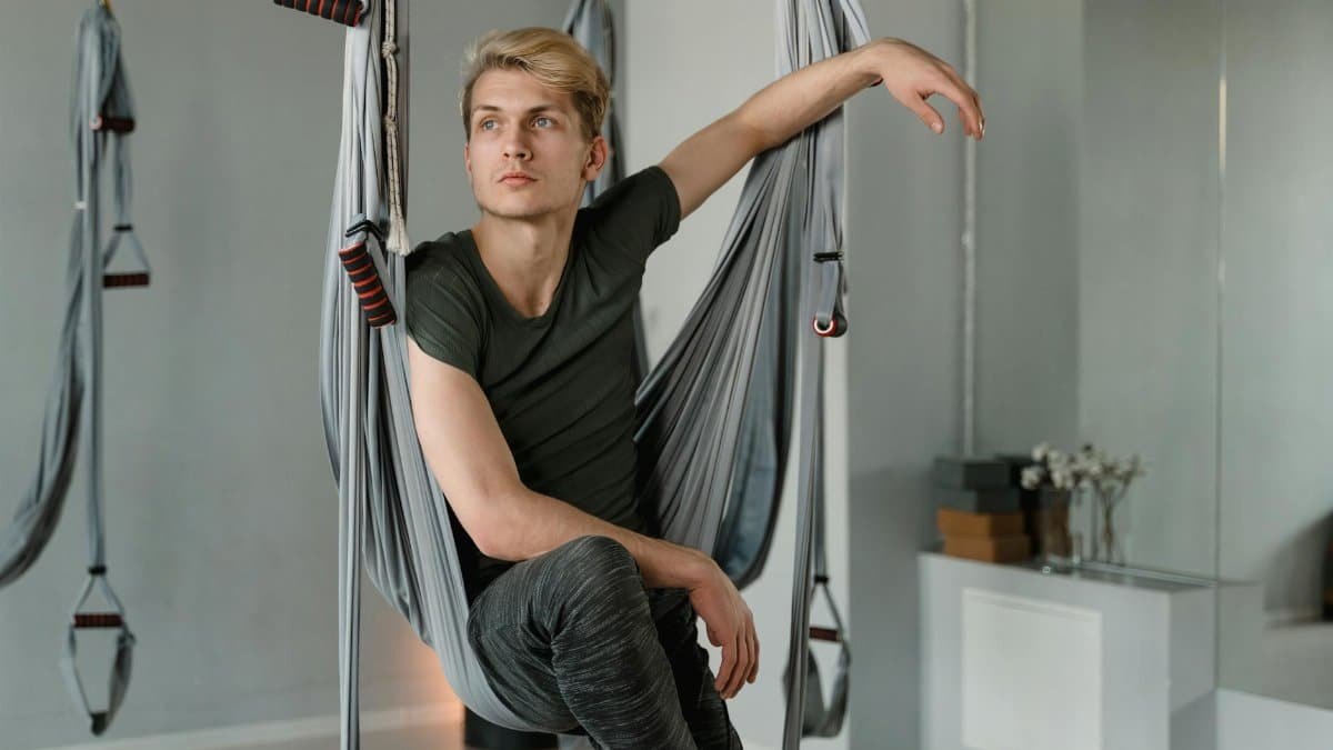 Adult man with blond hair in yoga studio using aerial swing for exercise.