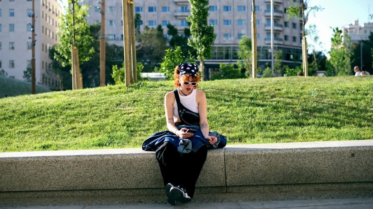 Redheaded woman in urban park, texting on phone with sunglasses, tattoos, and vibrant style.