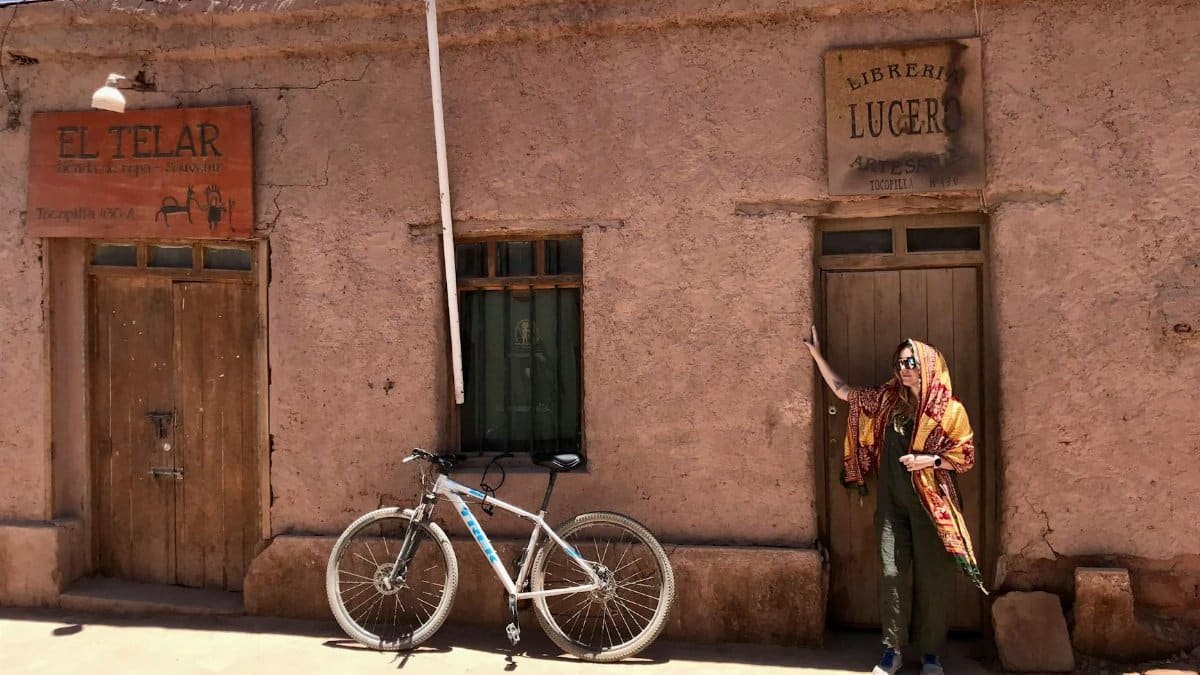 A person stands beside a rustic building with a bicycle under the bright sun.