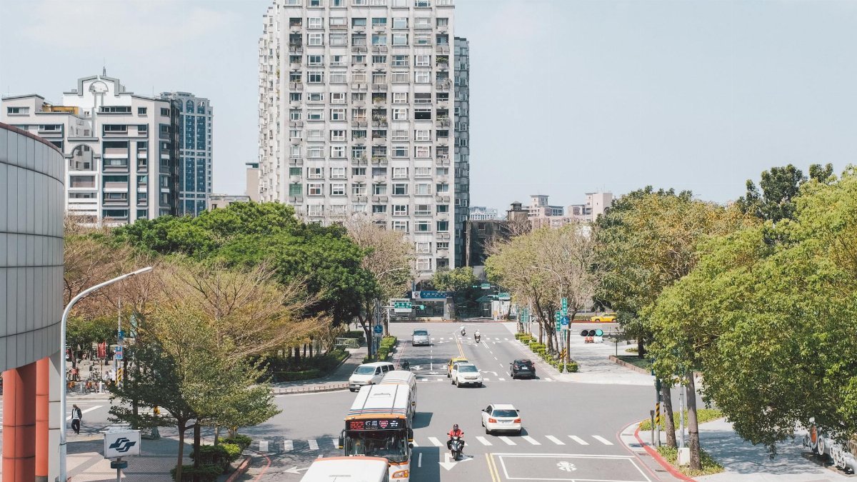 A busy city intersection with buses, cars, and a towering residential building under a clear summer sky.