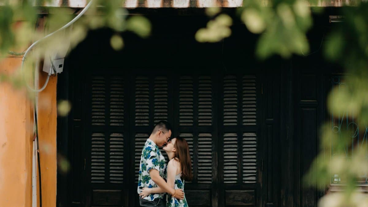 Side view of loving Asian couple hugging while standing near building in countryside with green leaves during date on summer day