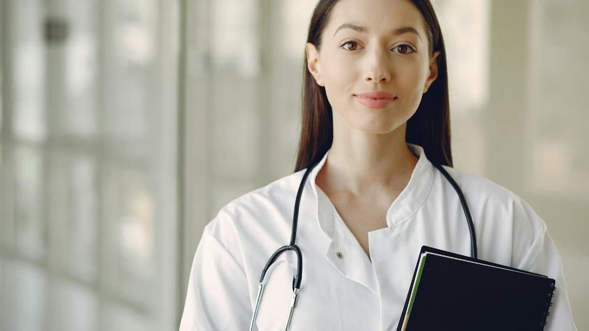 Crop smiling young ethnic female doctor in medical uniform with stethoscope and notebook standing in modern medical room with panoramic window