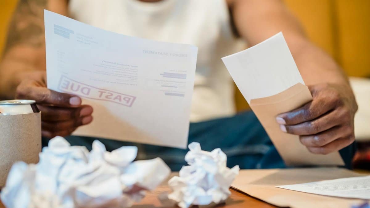 Man sitting indoors reviewing past due bills with crumpled papers on a coffee table.