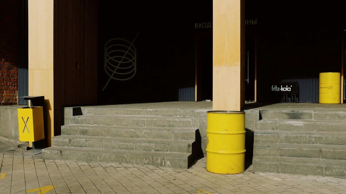 Exterior of modern building with concrete stairs located between columns and yellow barrel trash bins placed near wall on sunny day on street