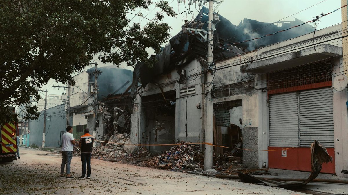 Two adults assess damage after an industrial warehouse fire. Smoke and debris visible.