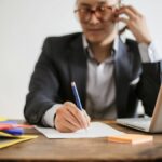 Businessman talking on phone while writing notes and using a laptop at desk.