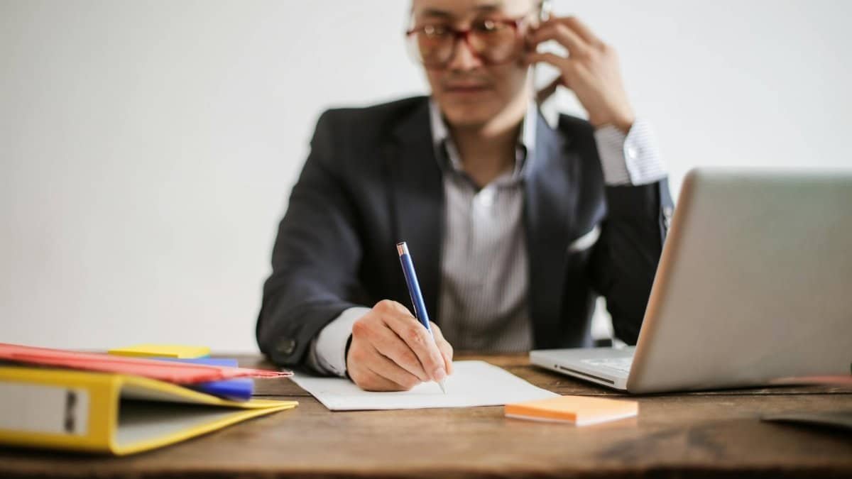 Businessman talking on phone while writing notes and using a laptop at desk.