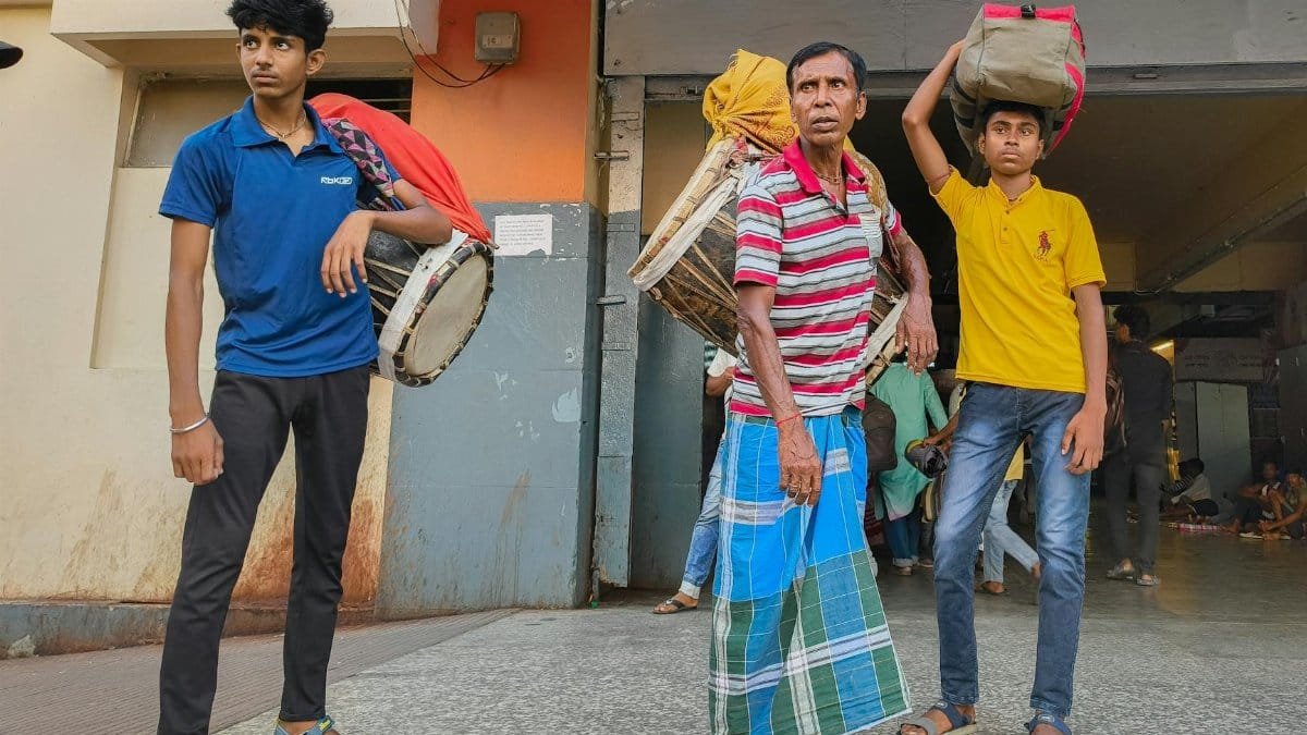 Three men carrying drums and bags in a public place. Captured outdoors.