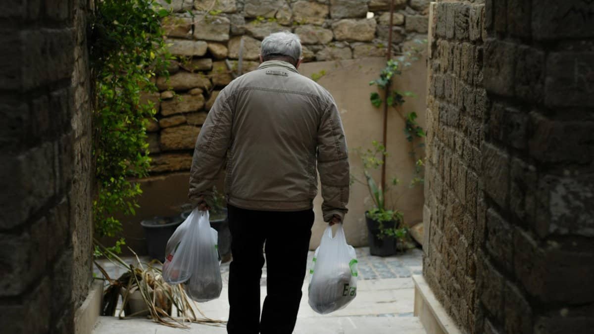 An elderly man carrying shopping bags walks through a narrow stone alley, providing a glimpse into daily life.
