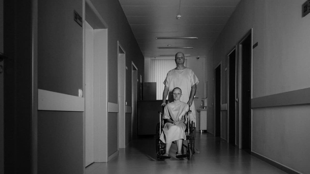 Two patients in hospital gowns, a man, and a wheelchair user, in a hospital hallway.