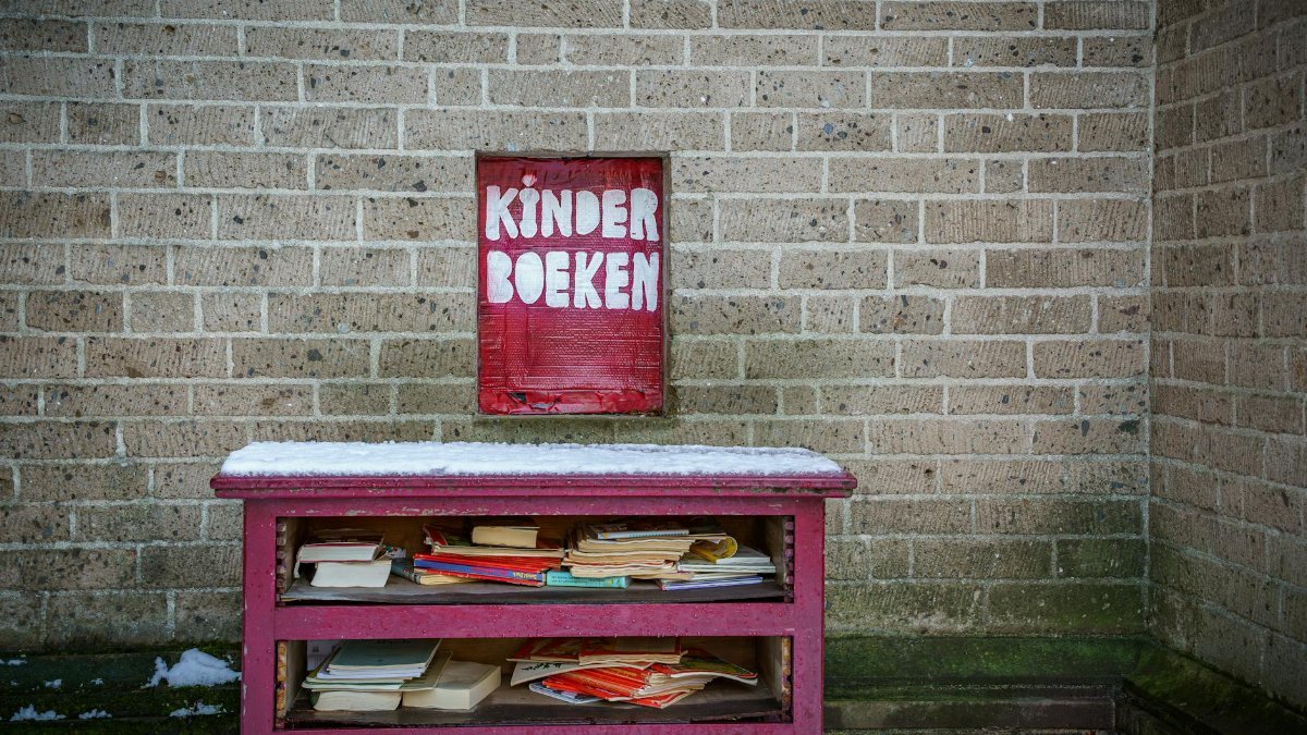 Red shelf with children's books marked 'Kinder Boeken' against a brick wall in winter.