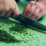 A person chopping fresh chives with a knife on a green cutting board, close-up view.