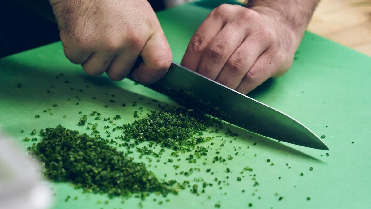 A person chopping fresh chives with a knife on a green cutting board, close-up view.