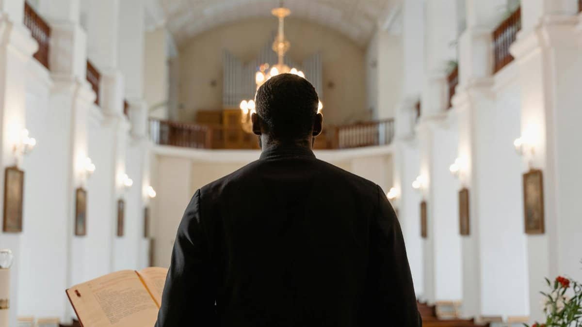 A priest stands before a congregation in a grand church, delivering a sermon with an open Bible.