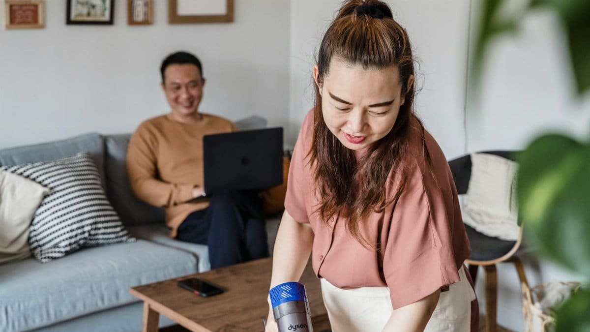 A woman smiling while vacuuming a living room, with a man working on a laptop in the background.