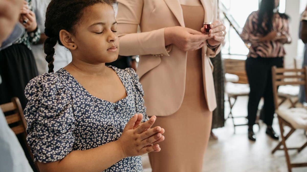 A young girl in a floral dress silently prays during a community gathering indoors.