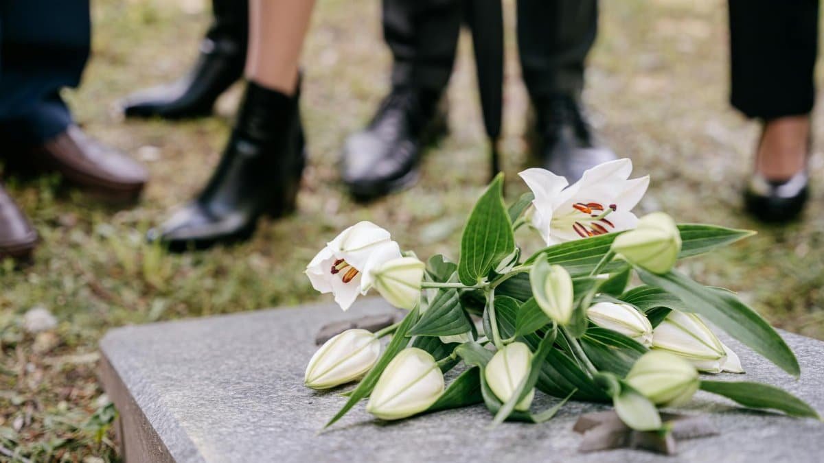 White lilies laid on a gravestone surrounded by people in mourning attire outdoors.
