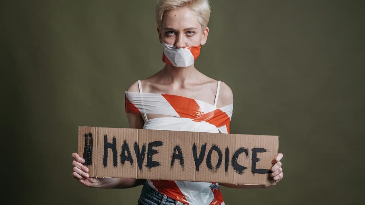 Woman wrapped in tape holding a protest sign 'I Have a Voice' in a studio setting.