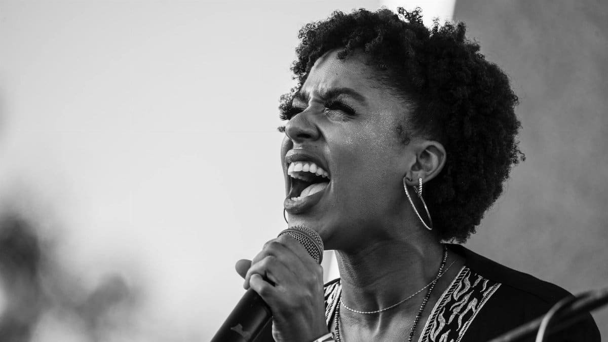 Black woman passionately speaking into a microphone outdoors. Grayscale close-up.