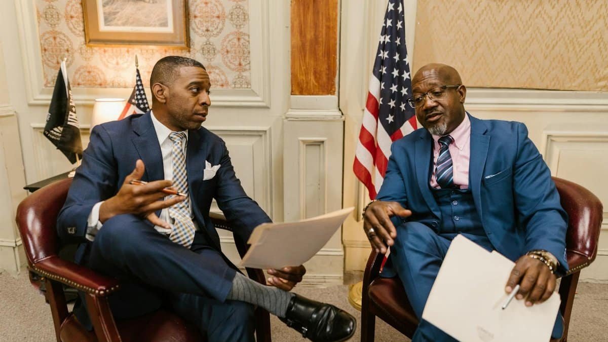 Two African American men in formal attire discussing documents in an office setting with flags in the background.