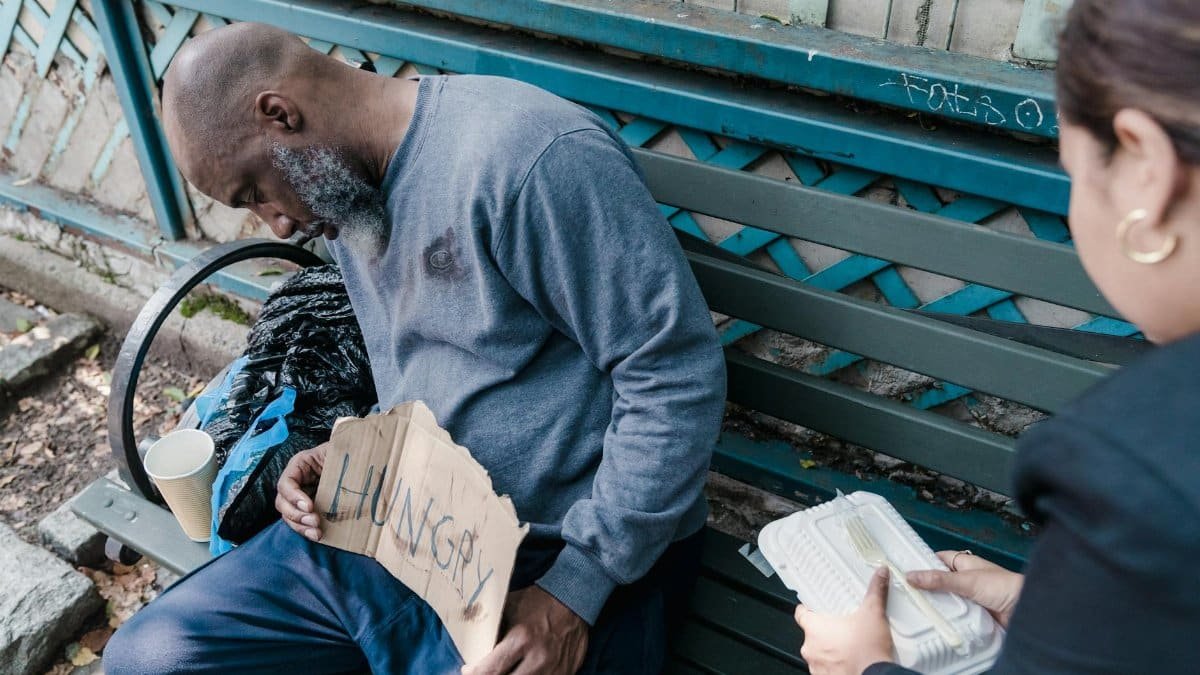 A woman compassionately offers food to a homeless man sleeping on a bench with a 'Hungry' sign.