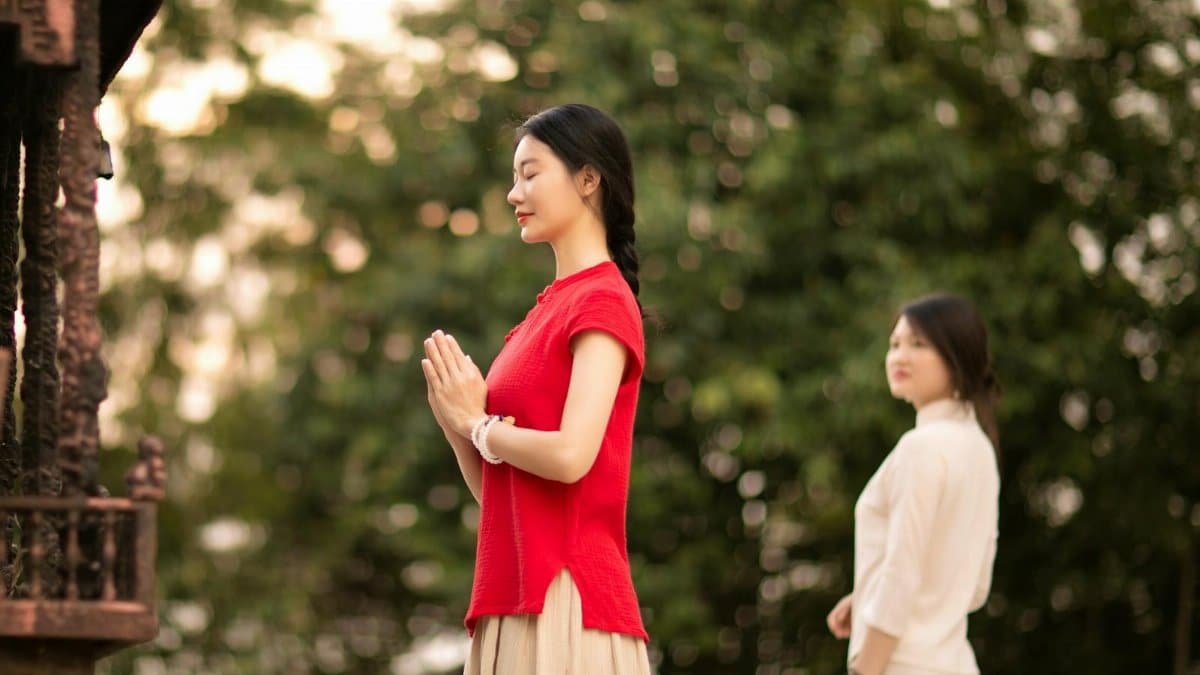 Serene image of two women meditating outdoors, one in red praying with eyes closed.