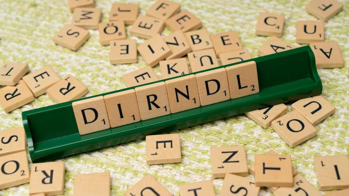 Scrabble tiles spelling 'dirndl' on a green tile holder with scattered letters on a textured surface.