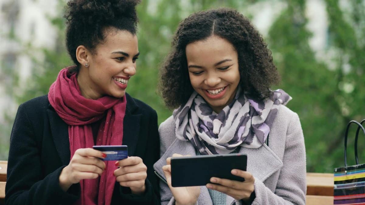 Two women happily shopping online with credit card and tablet outdoors.