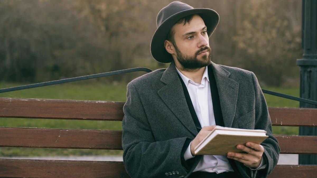 A bearded man in a hat sits pensively on a park bench, writing in a notebook. Captured in a serene outdoor setting.