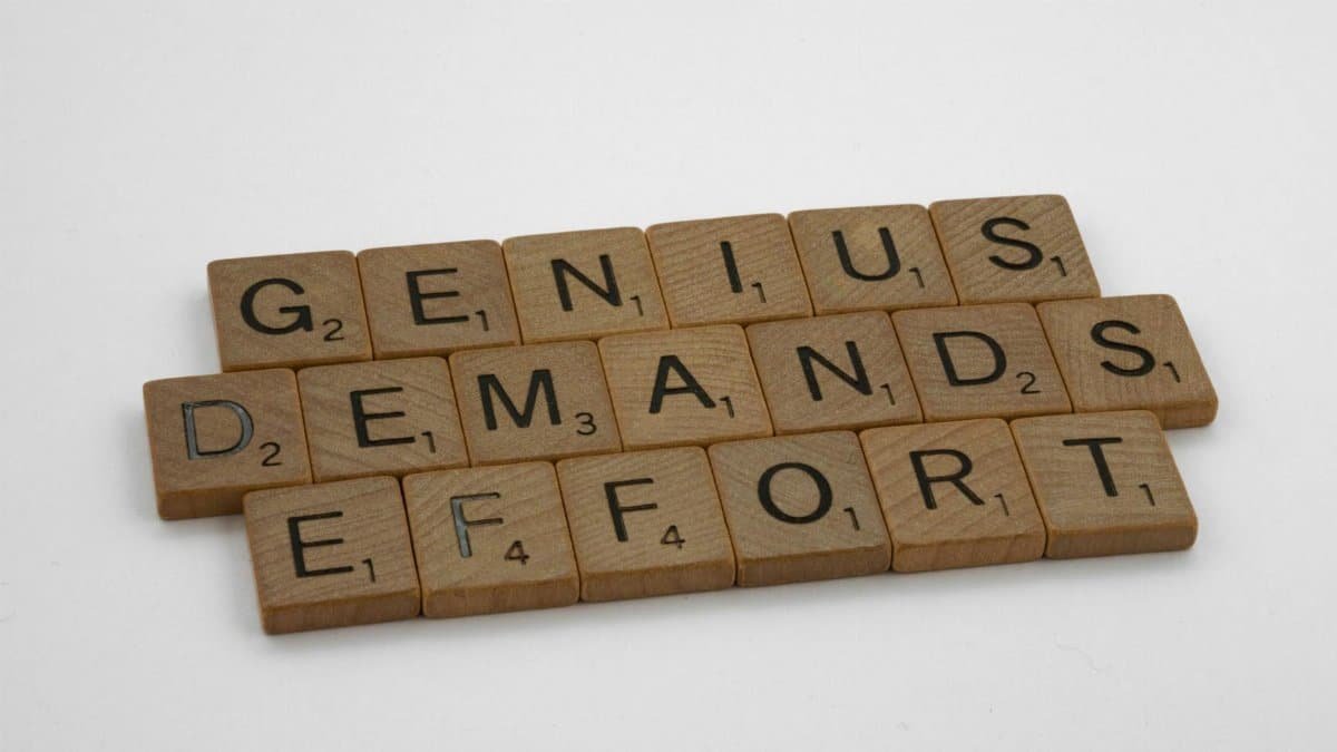 Scrabble tiles spelling 'Genius Demands Effort' on white surface, highlighting creativity and persistence.