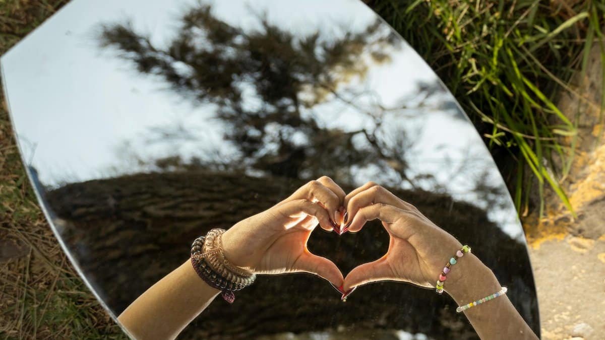 Reflection of hands forming a heart in a mirror outdoors, symbolizing love and connection.