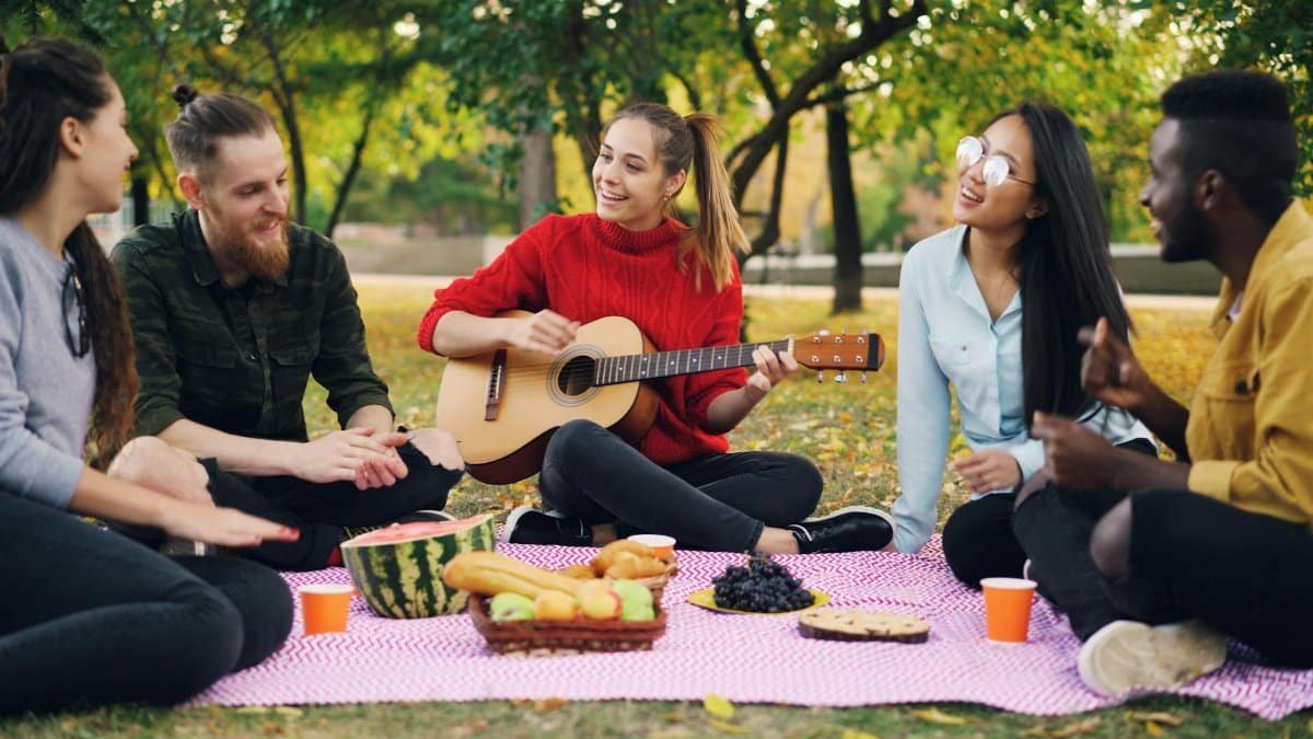 Young adults having a picnic with guitar music, fruit, and snacks in a park setting.