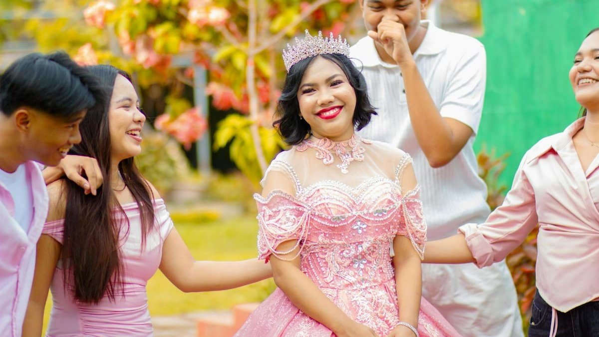 A young woman in a pink gown celebrates her quinceañera with friends in a bright, outdoor setting.