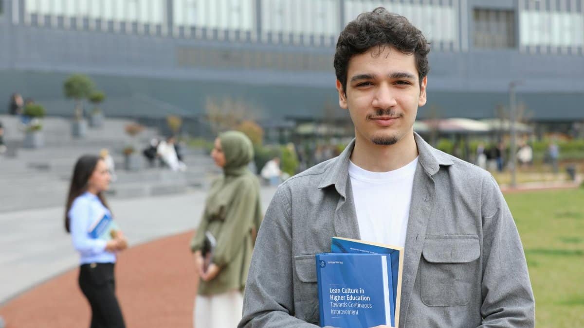 A young adult student stands outside on campus holding textbooks, symbolizing education.