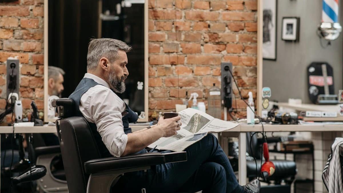 Middle-aged man sitting in a barbershop, holding a coffee and newspaper, surrounded by grooming tools.