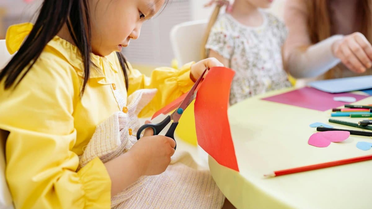 A young girl cuts colorful paper during an arts and crafts activity in a preschool classroom.