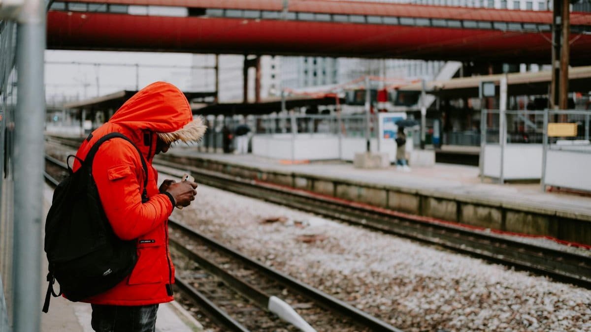 A man in a bright red jacket stands at a train station platform, checking his phone.