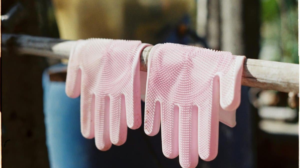 Two pink rubber cleaning gloves hanging to dry on a wooden stick outdoors in sunlight.