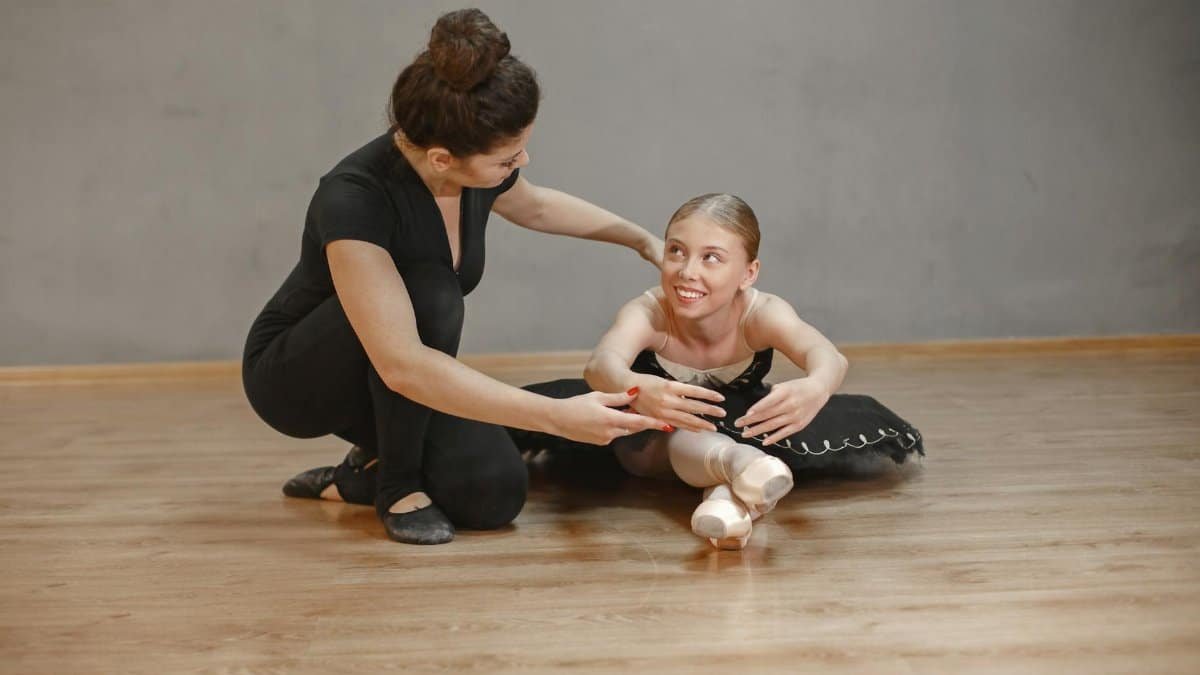 A ballet teacher assisting a young ballerina with stretching in a dance studio.