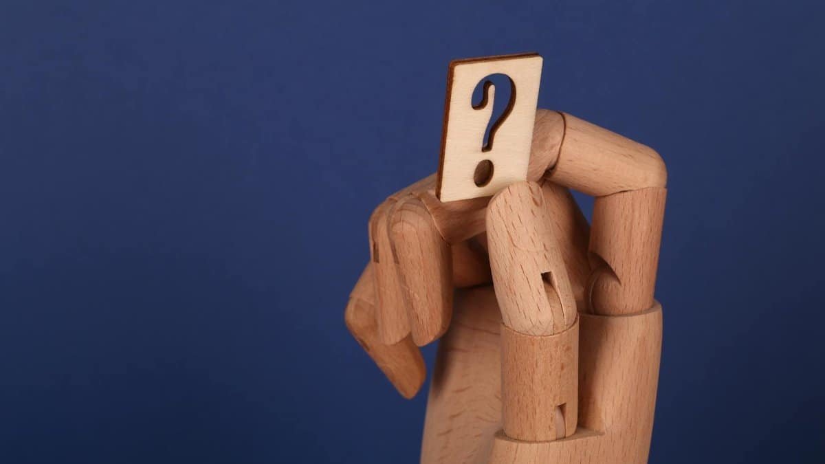 A wooden hand model holding a cut-out question mark against a blue background, symbolizing curiosity or inquiry.